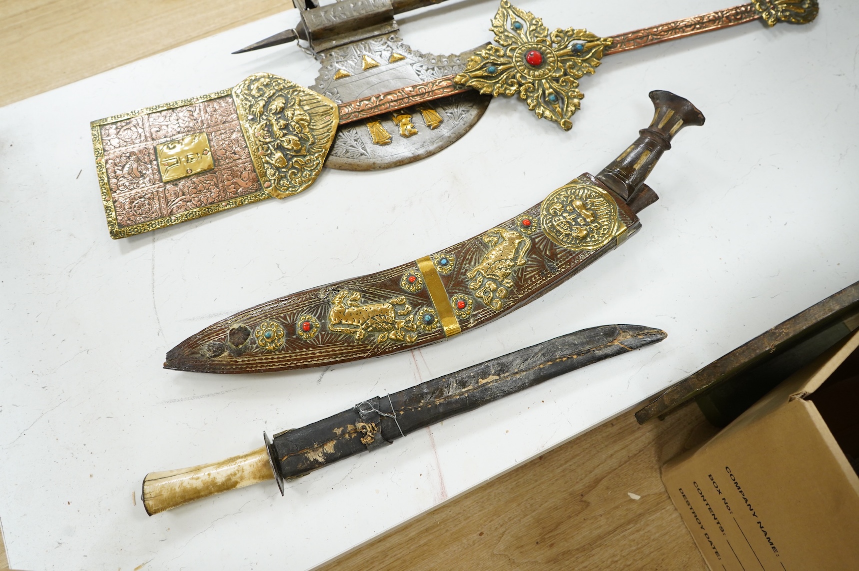 A decorative Indian axe, the large head inlaid with brass Hindu figures, together with a brass mounted North Indian kukri, another similar knife with bone handle, and a large shovel-shaped copper and brass item possibly